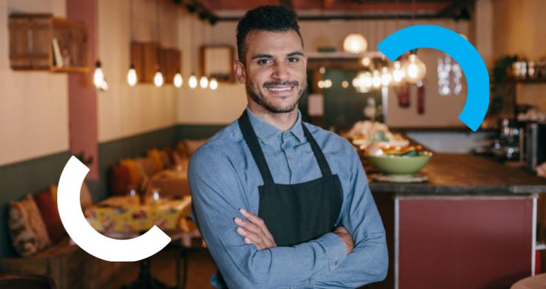 Pessoa de avental em pé com os braços cruzados no interior de um restaurante aconchegante.