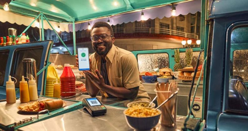 Uma pessoa no balcão de um food truck segurando um smartphone, com uma maquineta de cartão no balcão e rodeada de condimentos de comida de rua.