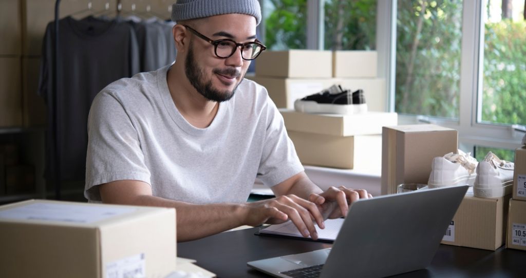 Empreendedor de e-commerce planejando ações de vendas em seu notebook, cercado por caixas de produtos, representando a aplicação de um plano de marketing para loja virtual.