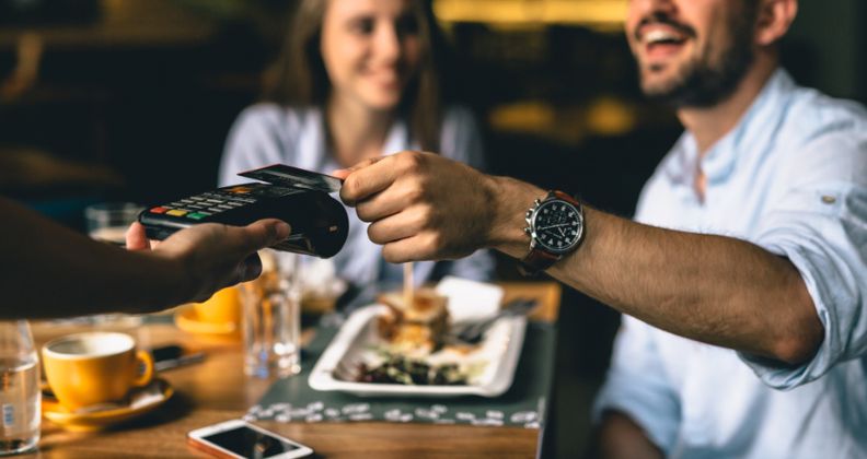 Pessoa pagando com cartão em um restaurante, com refeição e bebidas visíveis na mesa.