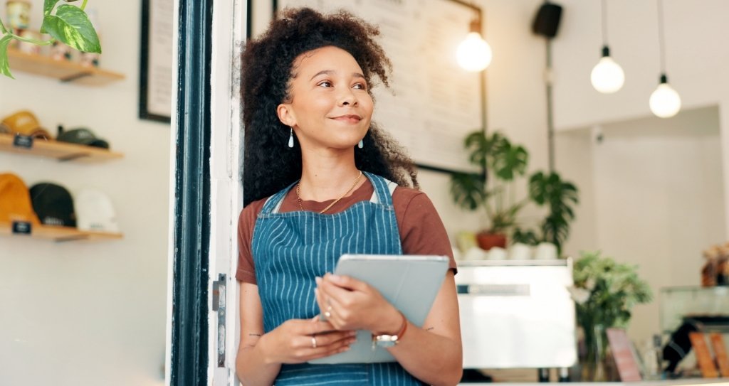 Empreendedora usando avental azul listrado segura um tablet na entrada de uma cafeteria iluminada, olha para o lado com expressão confiante, sugerindo planejamento financeiro e uso da antecipação de recebíveis para organizar o caixa.