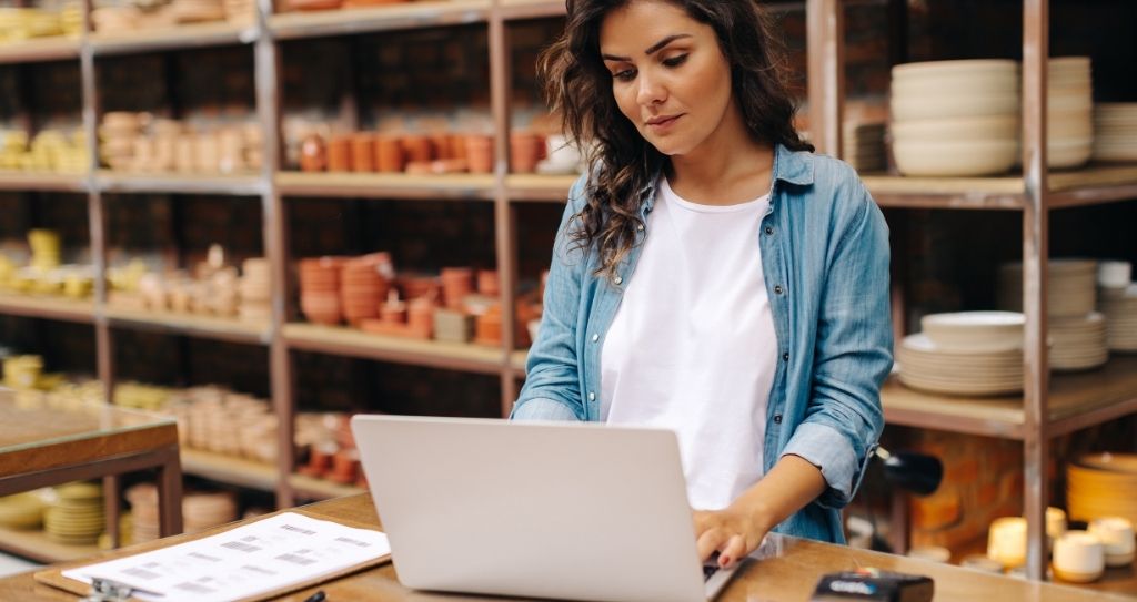 Uma mulher de cabelos castanhos ondulados, vestindo uma camisa jeans aberta sobre uma camiseta branca, trabalha concentrada em um notebook sobre um balcão de madeira. Ao fundo, prateleiras de madeira exibem diversos produtos de cerâmica e artesanato em uma loja bem iluminada, representando a rotina de uma empreendedora que analisa dados do negócio para identificar estratégias de aumento do ticket médio.