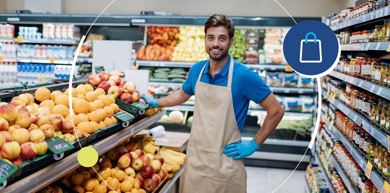 Empreendedor de avental faz pose em frente ao hortifruti do seu mercadinho de bairro.