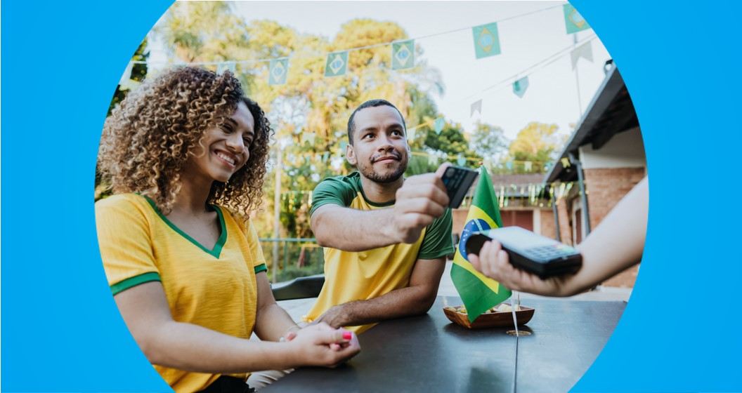 Um homem e uma mulher sentados em uma mesa de bar. O homem está com o braço estendido e aproximando o cartão da maquininha de pagamento. 
