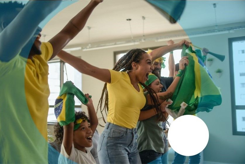 Seis pessoas pulando e comemorando os jogos do Brasil na Copa do mundo, em um ambiente decorado com bandeiras do Brasil.