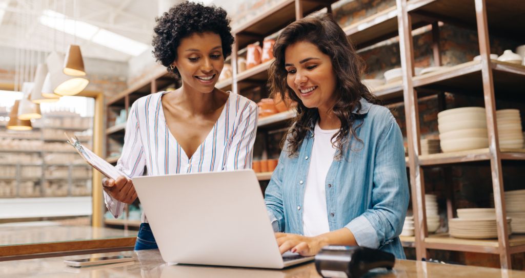 Duas empreendedoras analisam dados de vendas no notebook dentro de uma loja. Uma delas segura uma prancheta enquanto a outra digita, ambas sorrindo. A imagem representa o uso do Pix Parcelado e outras opções de parcelamento como ferramenta para organizar o caixa e impulsionar o negócio.