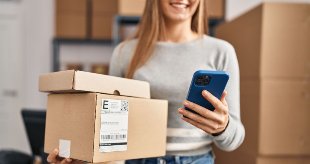 Alt: Mulher segurando caixas de devolução e usando o celular para acompanhar pedidos, representando a logística reversa e a praticidade das soluções para e-commerce Cielo.