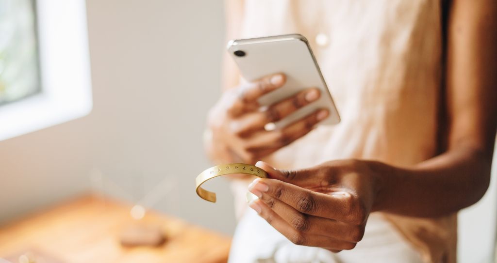 Pessoa segurando um smartphone em uma mão e uma pulseira dourada na outra, possivelmente tirando fotos para vender acessórios online.