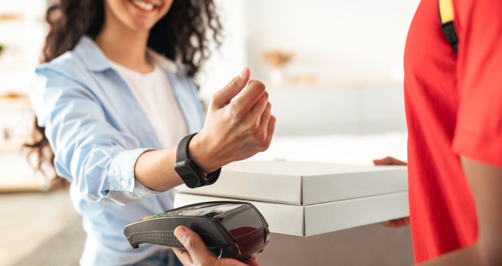 Mulher sorridente segura caixas de pizza e realiza pagamento com carteiras digitais, aproximando smartwatch (relógio inteligente) de maquininha.