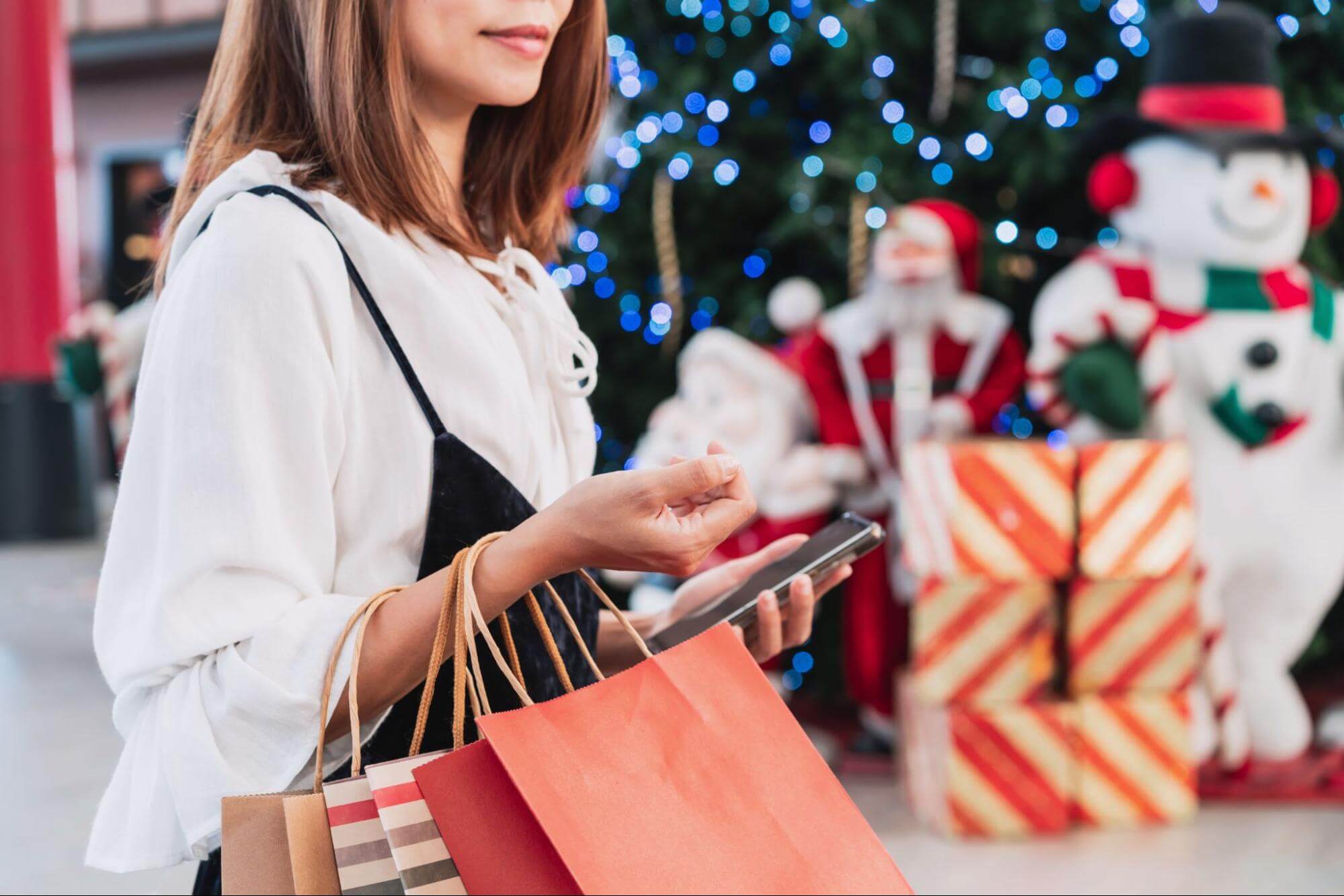 Mulher segurando sacolas de compras e usando o celular em um shopping decorado para o Natal, ilustrando como criar promoções de Natal para atrair clientes com experiências presenciais.
