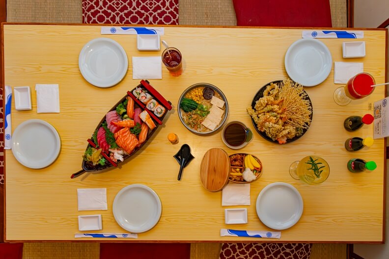 Mesa de restaurante com cinco pratos brancos e ao centro variadas comidas japonesas.