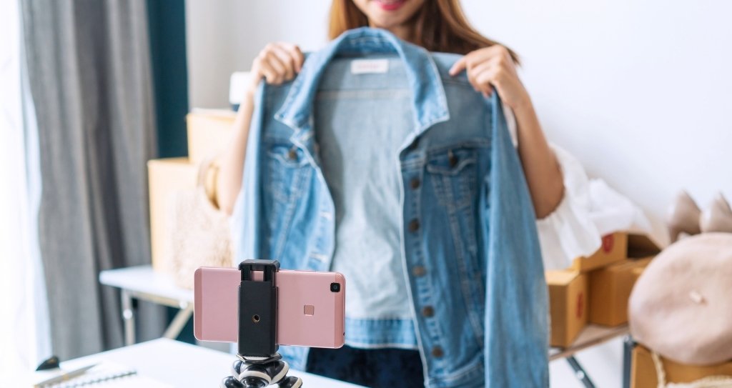 Empresária mostrando uma jaqueta durante uma live shop, interagindo com a audiência e promovendo o produto ao vivo, enquanto usa um smartphone para realizar a transmissão.