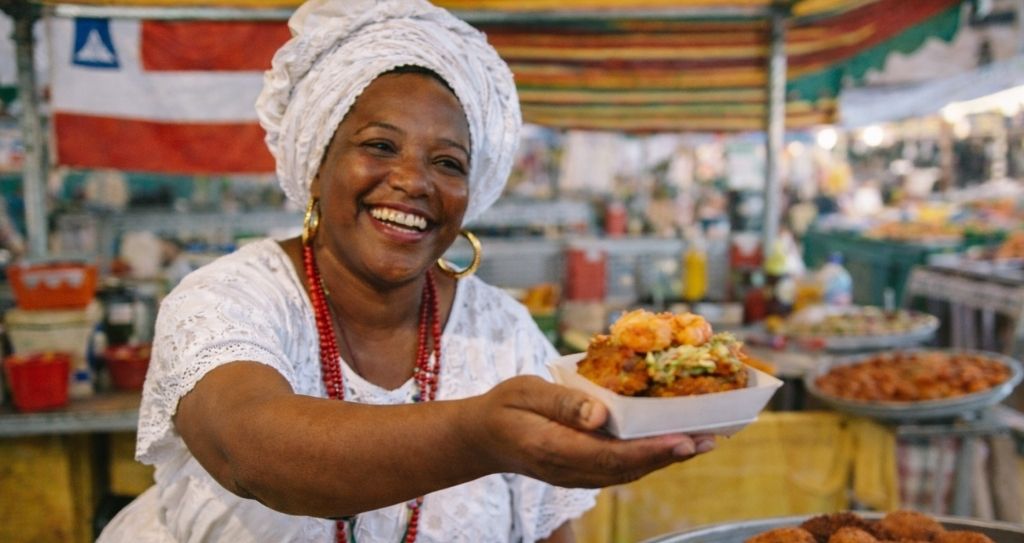 Uma mulher negra sorridente, vestindo roupas e turbante brancos tradicionais e colares de contas, oferece uma bandeja com um acarajé montado com camarões. A cena se passa em uma feira livre, com uma bandeira do estado da Bahia ao fundo da banca de acarajé. A imagem destaca a diversidade de opções de o que vender na feira no segmento de alimentação típica e regional.