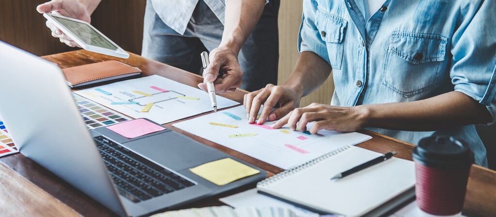 Duas pessoas sentadas em frente a um notebook gesticulando as mãos em cima de papéis e post-its, provavelmente debatendo alguma ideia. 