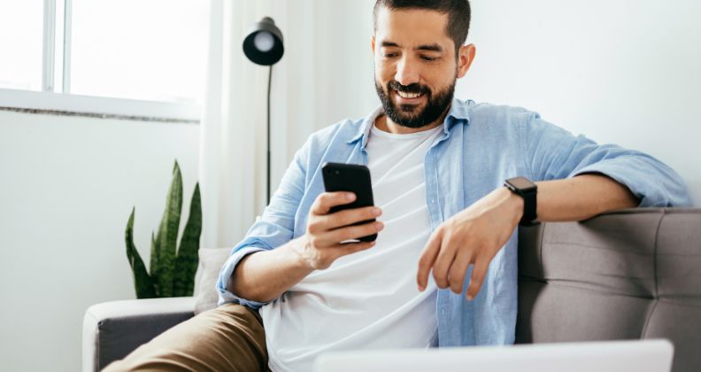 Empreendedor digital com uma camisa casual sentado em um sofá, segurando um smartphone com um laptop por perto. 