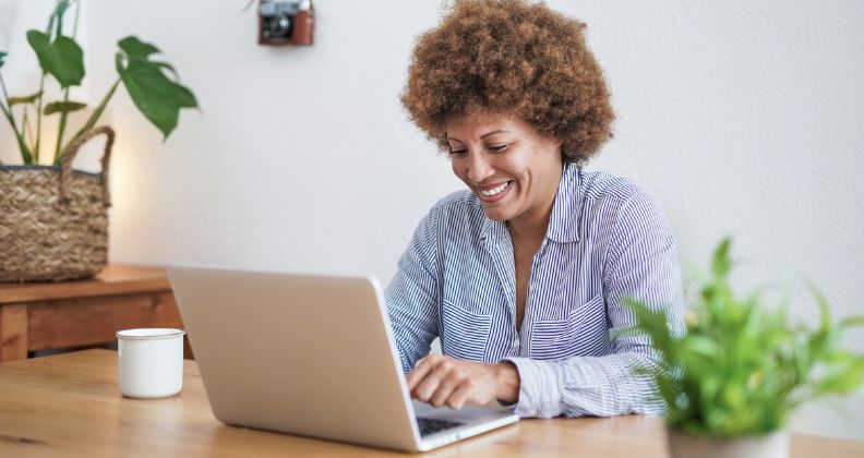 Empreendedora digital sentada em uma mesa com laptop e tablet, em uma sala com plantas e prateleiras.