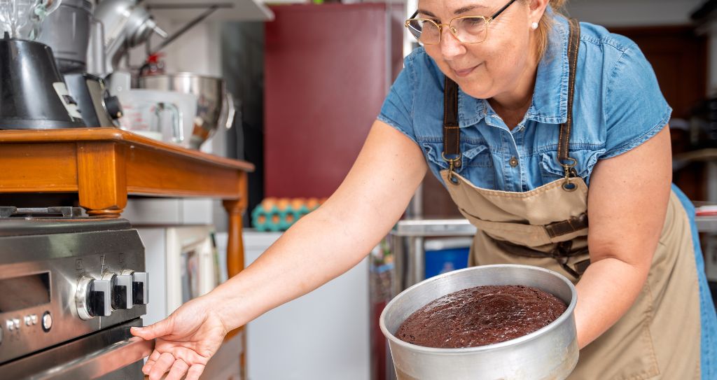 Confeiteira tirando bolo do forno em sua cozinha, ilustrando um exemplo de negócio lucrativo no ramo de doces e confeitaria artesanal.