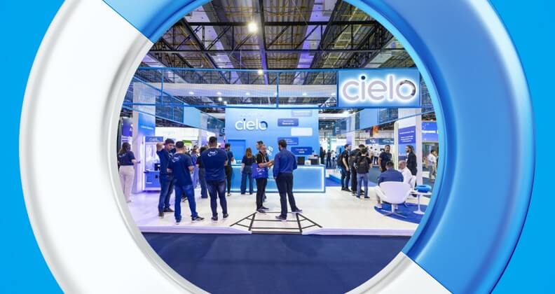 Fotografia do estande da Cielo na AUTOCOM 2025, mostrando a área de atendimento com colaboradores interagindo com visitantes. O espaço é iluminado, com painéis azuis, branding da Cielo visível e estrutura moderna voltada à demonstração de soluções tecnológicas para o varejo e meios de pagamento.