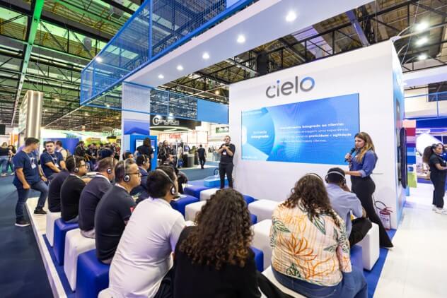 Foto do estande da Cielo na AUTOCOM 2025, onde um grupo de participantes está sentado, assistindo a uma apresentação do Cielo Talks enquanto dois profissionais falam ao público com apoio de uma tela grande exibindo conteúdos institucionais. O ambiente é moderno, bem iluminado e destaca o uso de tecnologia e inovação no ecossistema de meios de pagamento. 