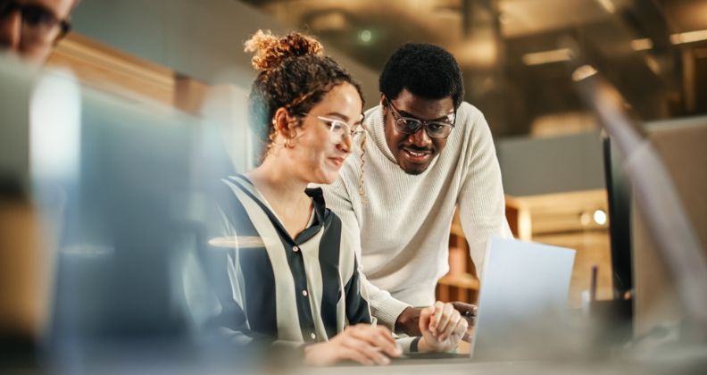 Dois colegas colaborando em uma mesa com um laptop e documentos, em um ambiente de escritório. 