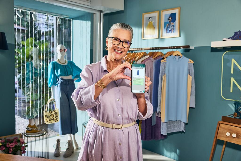 Senhora em uma loja de roupas segurando um celular, como se estivesse mostrando uma nova funcionalidade no celular dela. 