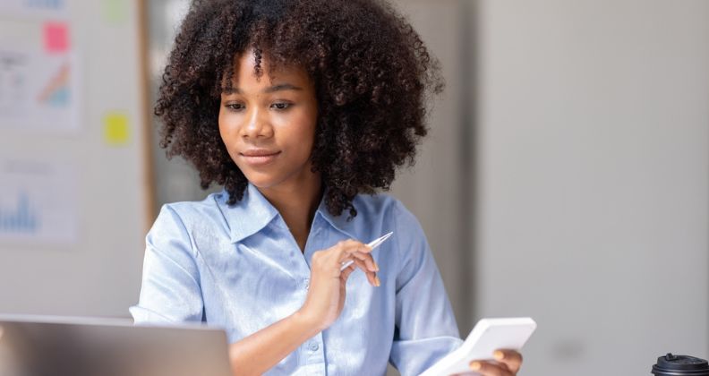 Mulher de camisa azul segurando caneta e smartphone em uma mesa com laptop e xícara de café.