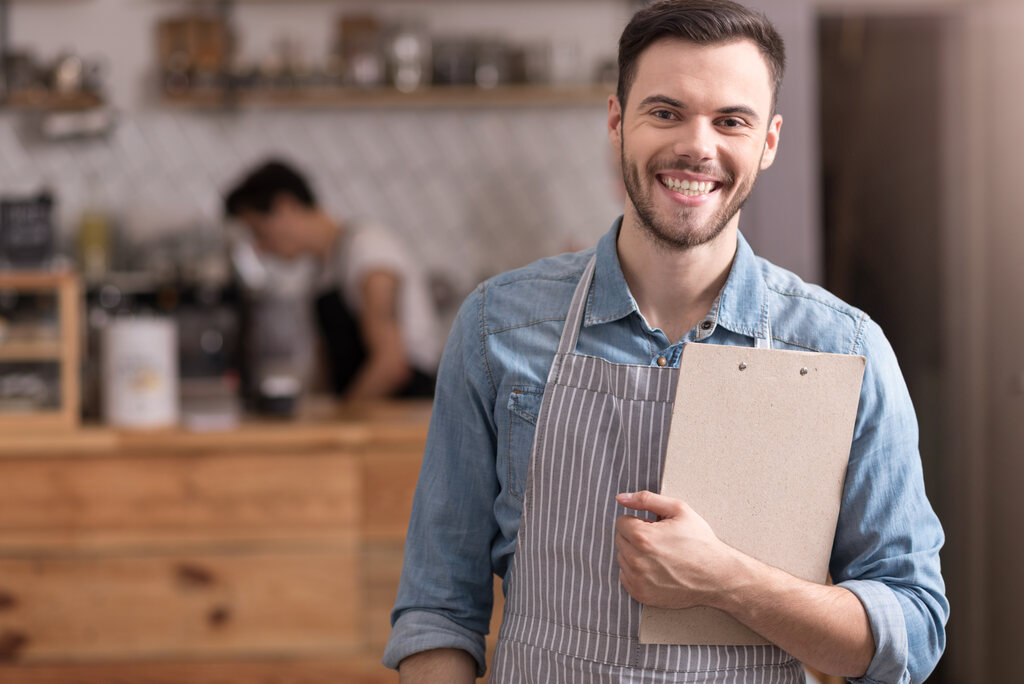 Homem vestido com um avental e segurando uma prancheta em um bar, provavelmente ele está empreendendo no ramo de bebidas. 