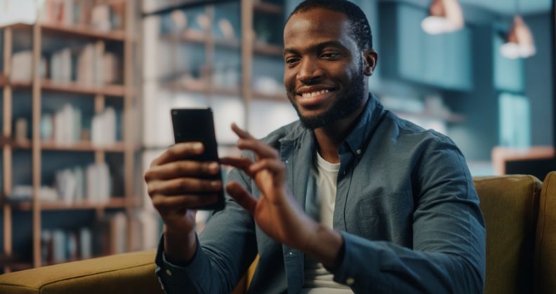 Homem de camisa azul sentado em um sofá amarelo segurando um smartphone em uma sala com estantes.