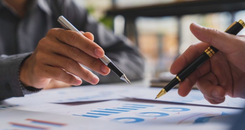 Duas pessoas fazendo análise financeira com gráficos e tabelas em papel com canetas na mão.