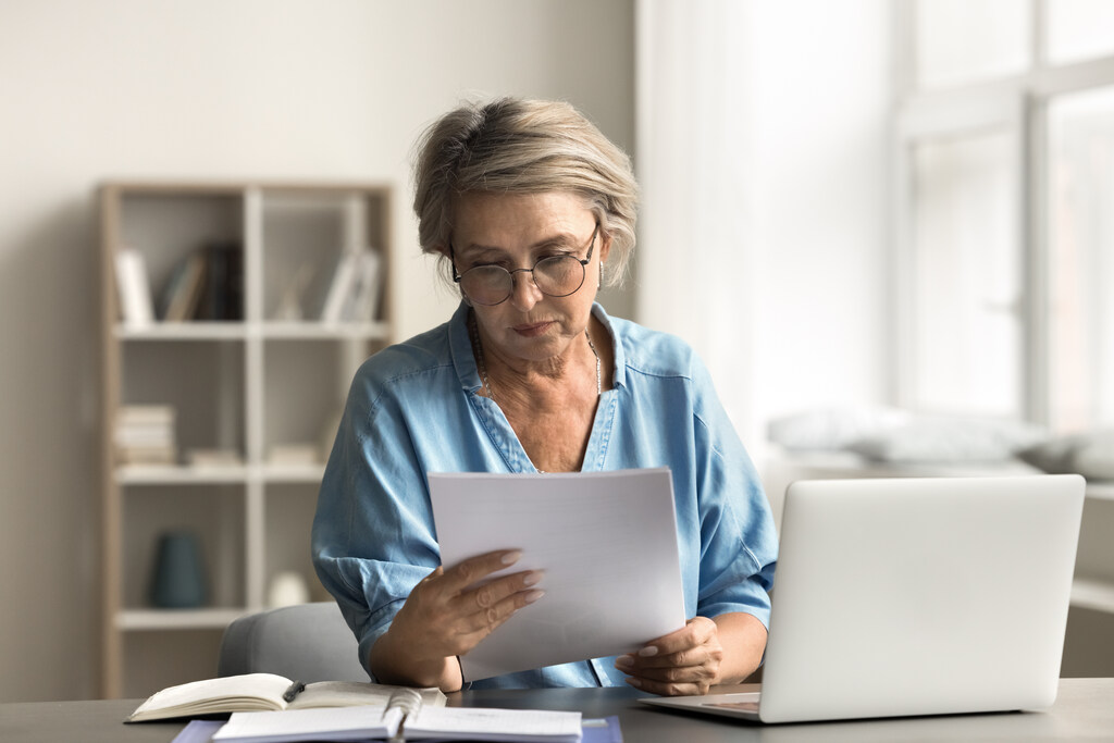 Mulher sentada em frente a um notebook e segurando em uma das mãos algum tipo de documento, provavelmente está checando se o documento está correto. 