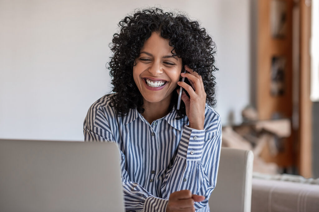 Mulher sorridente ao telefone sentada em uma mesa de frente para um notebook, provavelmente atendendo uma ligação que a deixou feliz. 