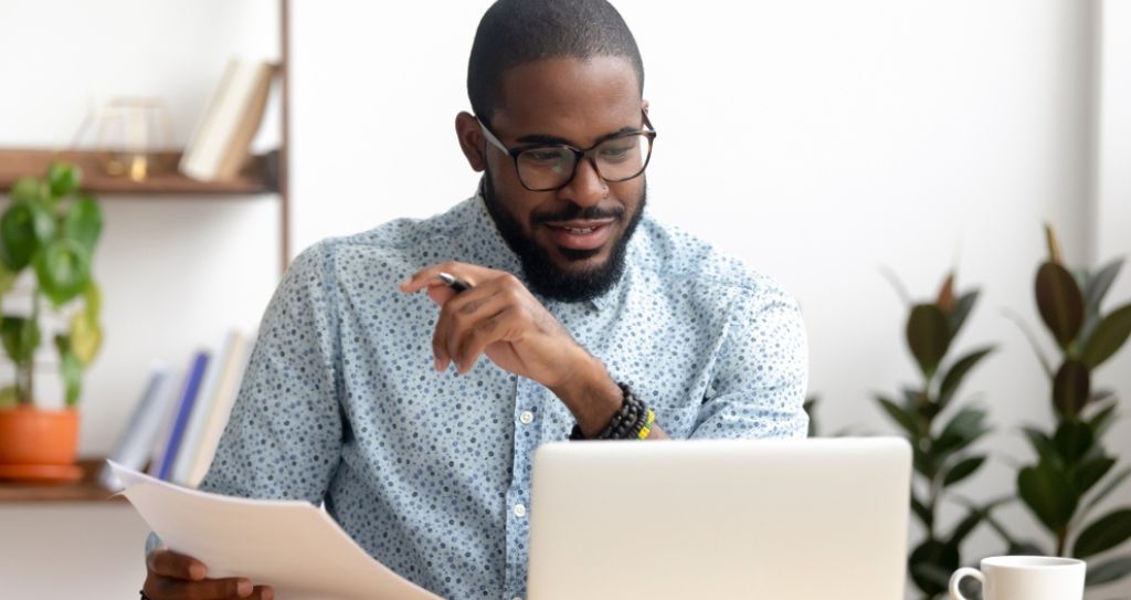 Pessoa com camisa estampada segurando um documento em uma mesa com um laptop e uma xícara de café.