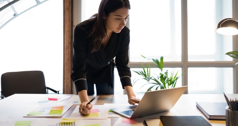 Pessoa em pé em frente a uma mesa com laptop, papéis e post-its em um escritório bem iluminado analisando indicadores financeiros.