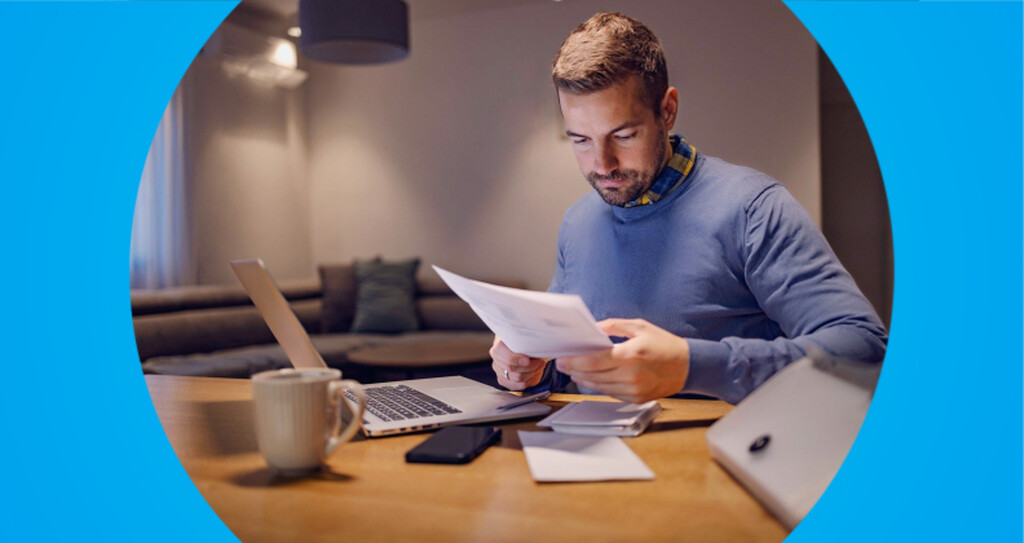 Homem sentado de frente a um notebook e segurando com as mãos um documento, como se estivesse conferindo o que está escrito.