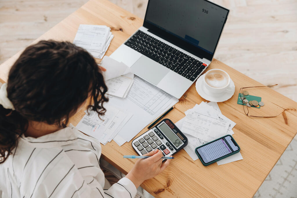 Imagem área de uma mulher sentada em frente a um notebook com vários papéis e uma calculadora e caneta na mão, possivelmente calculando alguma taxa.