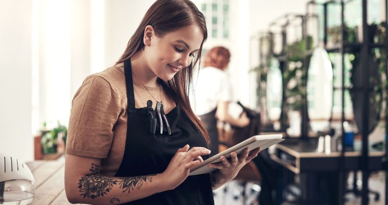 Mulher de braços tatuados com avental segura um tablet em um salão de beleza.