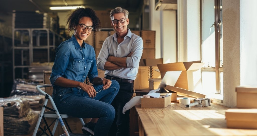 Duas pessoas empreendedoras, sorrindo, em um ambiente de estoque e embalagens, representando pequenos negócios organizando seus envios. A imagem reforça o contexto de quem já vende online e precisa de um checkout de pagamento eficiente e confiável para profissionalizar ainda mais as vendas.