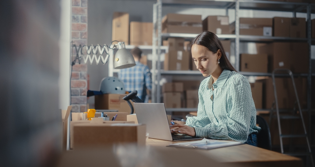 Pessoa empreendedora utilizando um notebook em meio a caixas de pedidos no estoque de um e-commerce. A imagem ilustra a pessoa fazendo pesquisas para escolher um checkout de pagamento otimizado para facilitar o dia a dia das vendas pela internet.