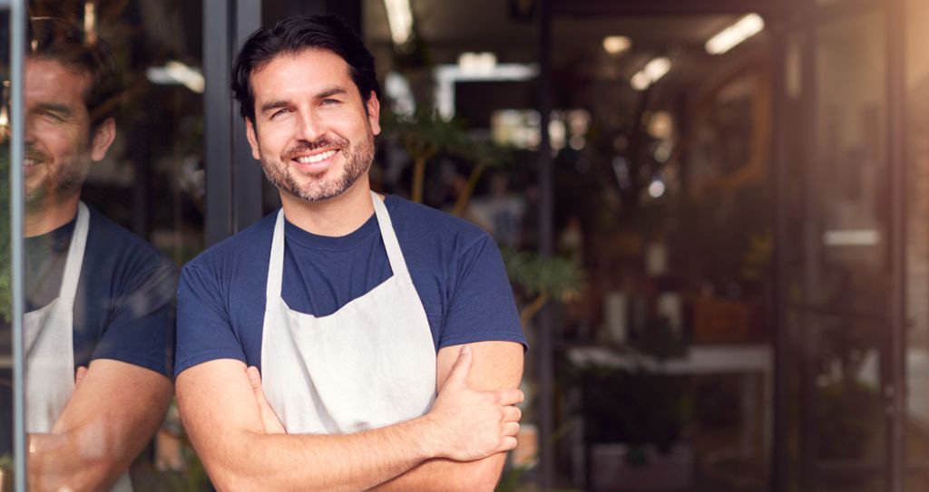 Empreendedor ME ou EPP de avental com braços cruzados em frente a uma loja.