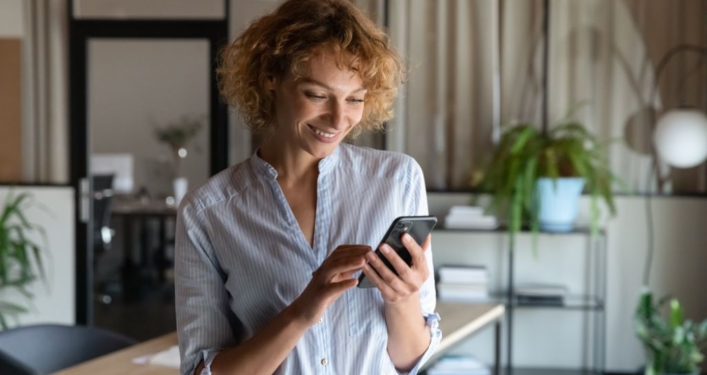 Mulher sorridente, de cabelos cacheados e curtos, usando um smartphone em um ambiente de trabalho moderno fazendo estratégias omnichannel, com plantas e prateleiras ao fundo.