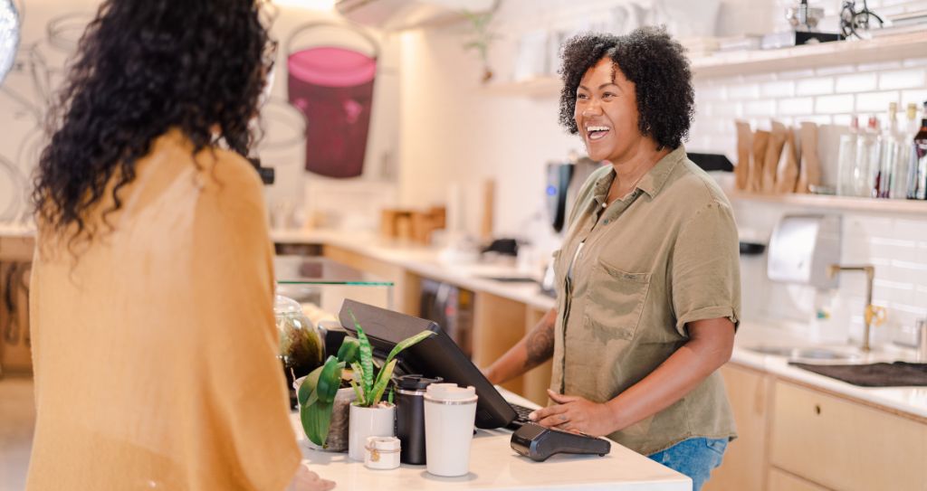 Duas mulheres no balcão de um café, uma ME ou EPP trabalhando na caixa registadora e outra de costas.