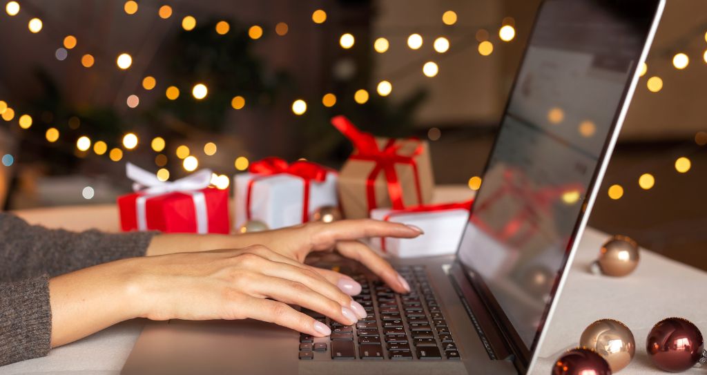 Close-up de mãos digitando em um notebook, com uma árvore de Natal desfocada ao fundo, representando a busca online por ideias do que vender no Natal. 