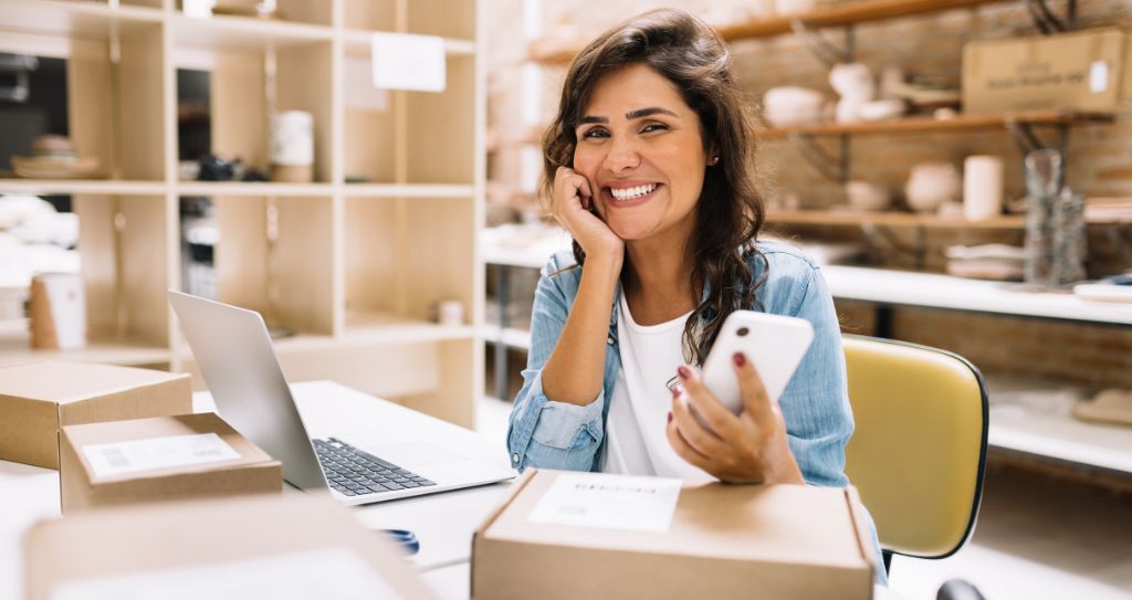 Pessoa sentada em uma mesa entre caixas de papelão, com laptop e smartphone, enquanto sorri depois de ter configurado split de pagamento.