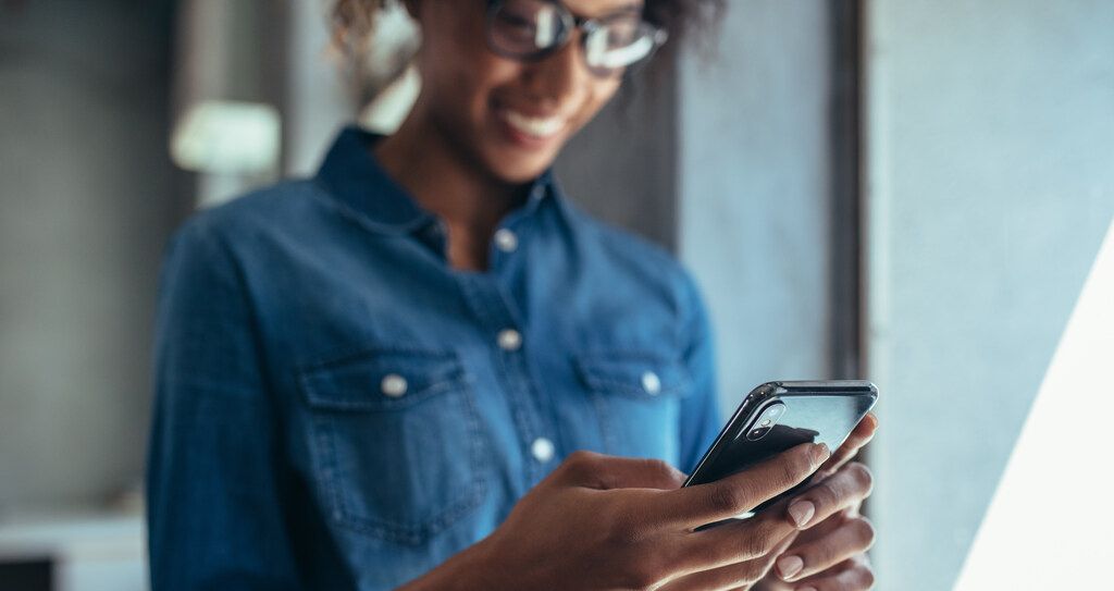 Mulher sorrindo enquanto usa o celular, representando o consumo de infoprodutos como cursos online, e-books ou podcasts diretamente pelo smartphone.