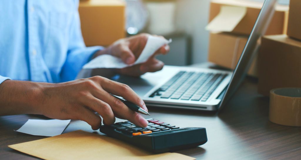 Pessoa calculando custos e lucros com produtos para revenda, utilizando notebook e calculadora em ambiente com caixas de envio.