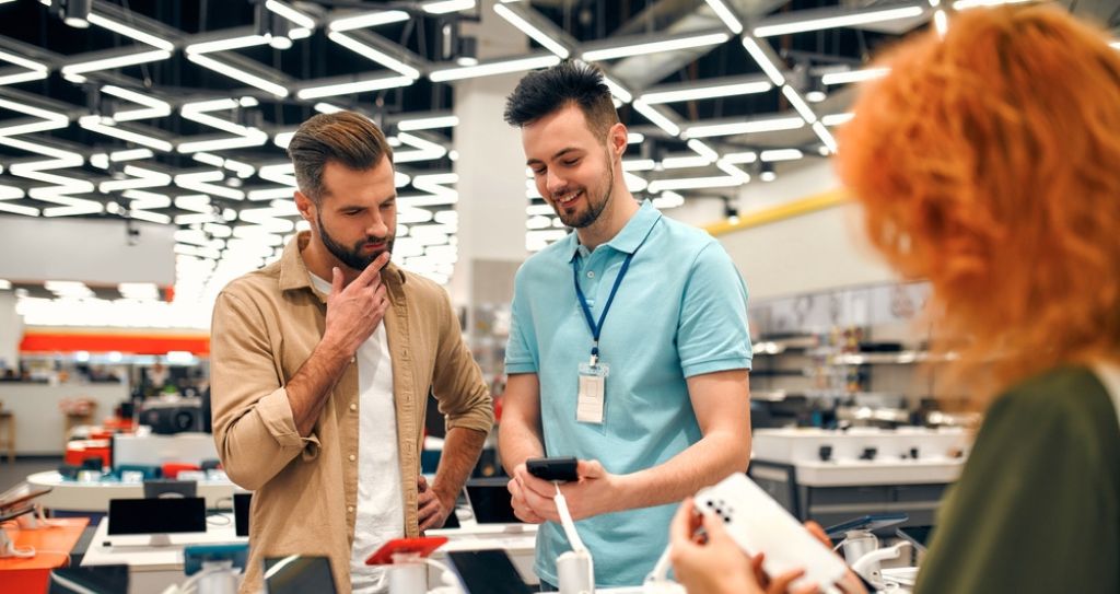Vendedor apresenta smartphones em loja física, um dos itens mais vendidos na Black Friday. O cliente ouve o vendedor enquanto uma mulher analisa outro modelo.