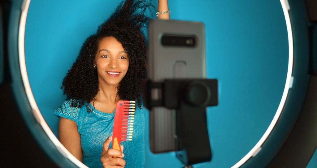 Uma criadora de conteúdo sorridente com cabelos cacheados se posiciona em frente a um ring light e um smartphone para gravar um vídeo de demonstração. Ela segura um pente colorido e interage com a câmera, exemplificando o uso de acessórios de iluminação e suporte que são excelentes produtos para vender na internet para o público que trabalha com redes sociais e produção de vídeo. O fundo azul vibrante destaca a cena e reforça o aspecto profissional da produção.