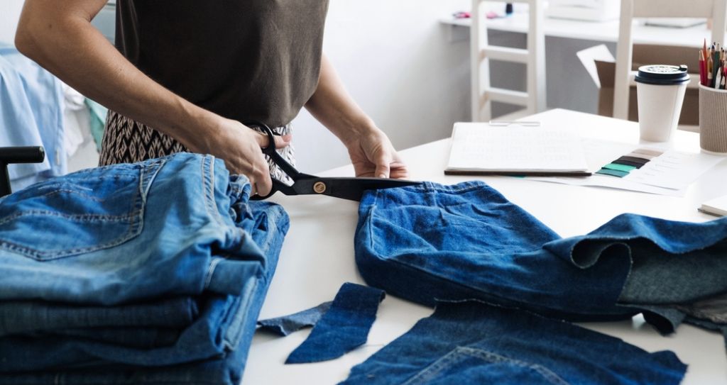 Pessoa cortando calça jeans para reaproveitamento em artesanato, mostrando uma ideia prática de como fazer renda extra com customização de roupas usadas e dar novo valor ao que já não é utilizado.