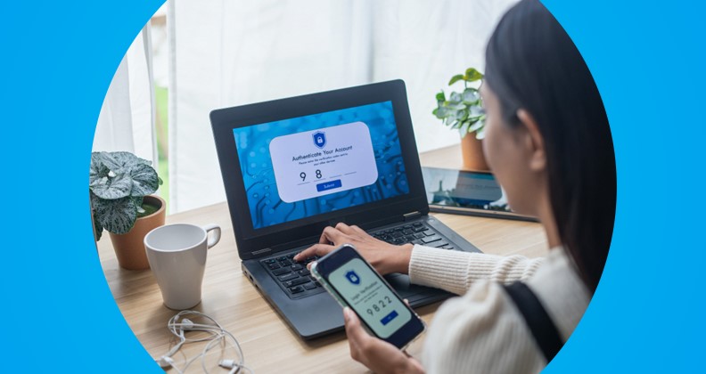 Pessoa usando autenticação em dois fatores para acessar conta em laptop, digitando código exibido no smartphone. Na tela do computador aparece um formulário de verificação com campo para inserir código e botão 'Submit'. Ambiente com mesa de madeira, plantas, caneca branca e fones de ouvido.