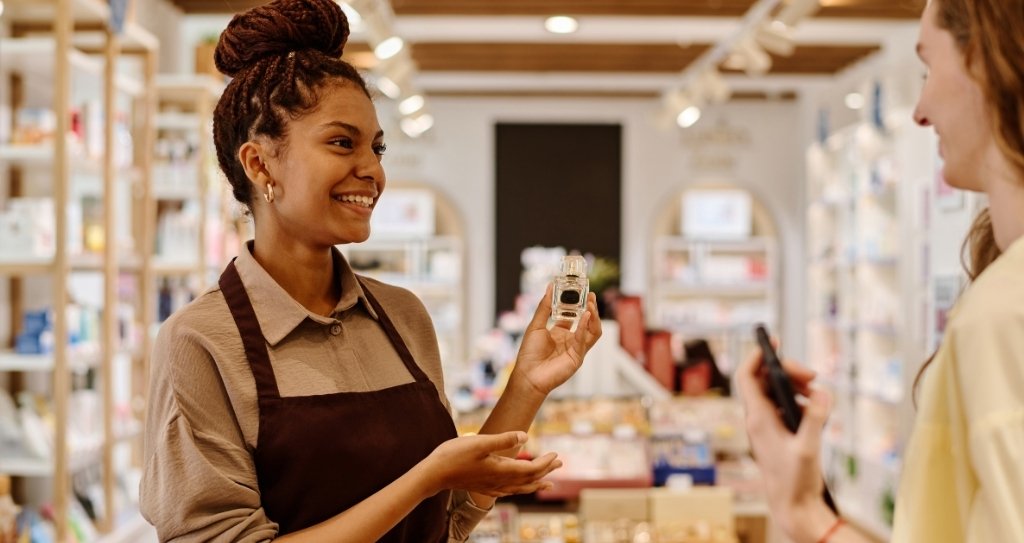 Vendedora em loja física mostrando um perfume para cliente, exemplificando a etapa de prospecção do ciclo de vendas, onde a interação inicial é feita para atrair o cliente e despertar seu interesse no produto.
